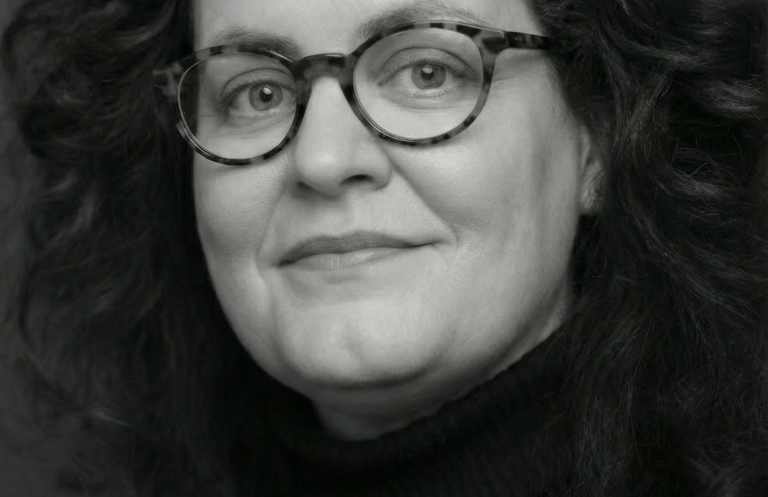 Cornelia Schmitz - Führungserfahrung als Abteilungsleiterin und Talentförderin. Porträt einer Frau mit lockigem Haar und Brille, lächelnd vor neutralem Hintergrund.
