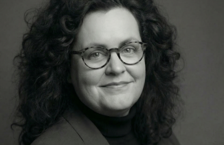 Cornelia Schmitz - Mehr als 40 Jahre Berufserfahrung. 10 Jahre international tätig gewesen. Porträt einer lächelnden Frau mit Brille und lockigem Haar auf dunklem Hintergrund.