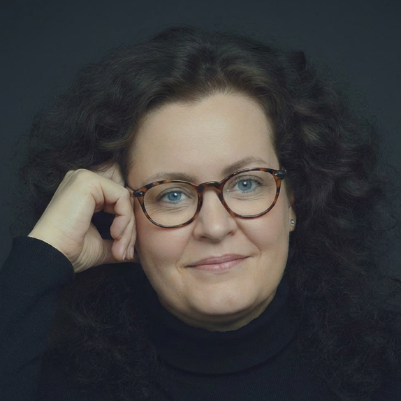 Porträt von Cornelia Schmitz, einer Frau mit lockigem, dunklem Haar und Brille. Sie blickt freundlich und aufgeschlossen.