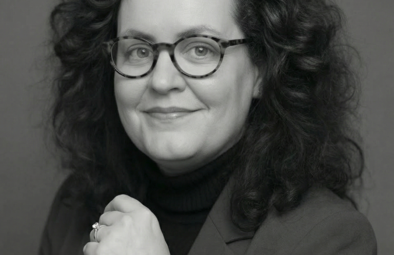 Cornelia Schmitz - Erfolge als Projektleiterin und Konzeptentwicklerin. Porträt einer lächelnden Frau mit lockigem Haar und Brille vor einem dunklen Hintergrund.