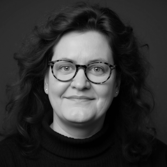Cornelia Schmitz - Die Mentorin für Führungskräfte. Porträt von Cornelia Schmitz, einer Frau mit lockigem, dunklem Haar und Brille. Sie blickt freundlich und aufgeschlossen.