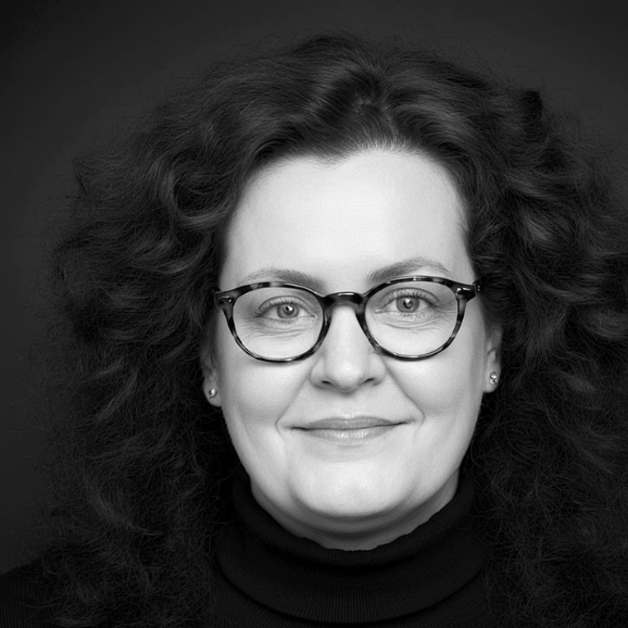 Porträt von Cornelia Schmitz. Eine Frau mit lockigem Haar und Brille, freundlich lächelnd und mit souveränem Blick.
