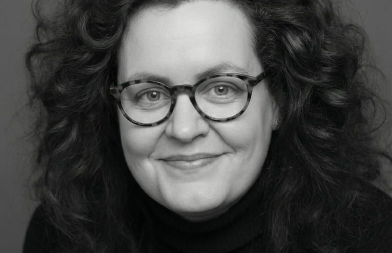Cornelia Schmitz - Von der Mitarbeiterin zur Führungskraft. Lächelnde Frau mit lockigem, schwarzem Haar und Brille, in dunkler Kleidung vor neutralem Hintergrund.