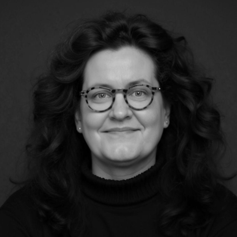 Porträt von Cornelia Schmitz, einer lächelnden Frau mit lockigem, dunklem Haar und Brille. Sie blickt freundlich und aufgeschlossen.