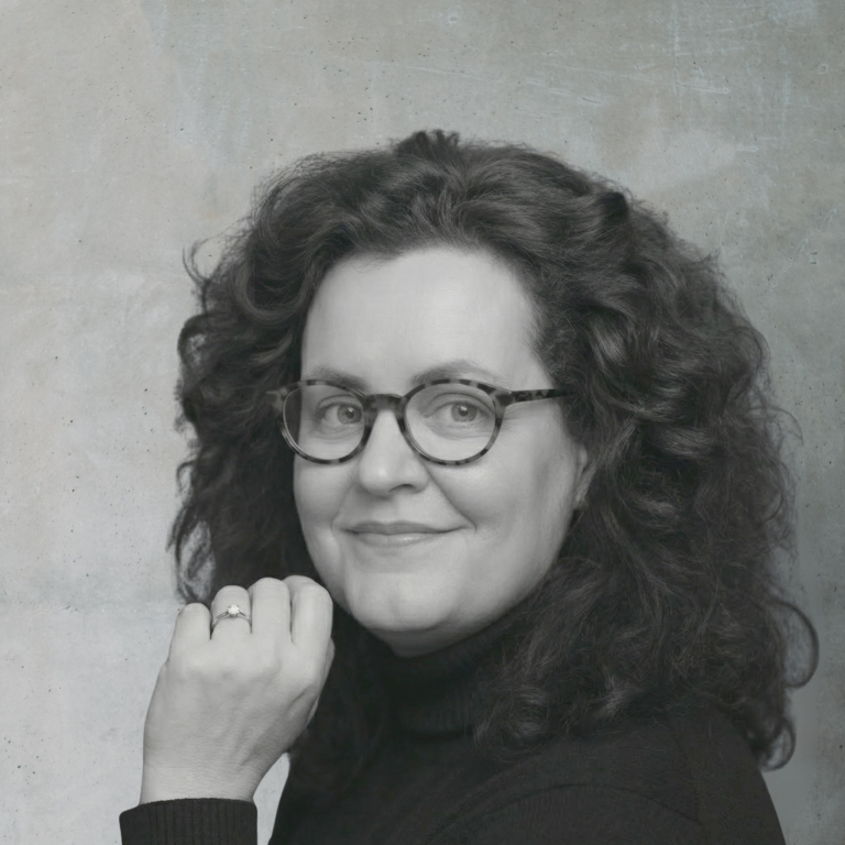 Cornelia Schmitz - Mentorin für Führungskräfte und Co-Host bei 'Sinnstiftendes Unternehmertum'. Porträt von Cornelia Schmitz, einer Frau mit lockigem, schulterlangem Haar und Brille, freundlich lächelnd.