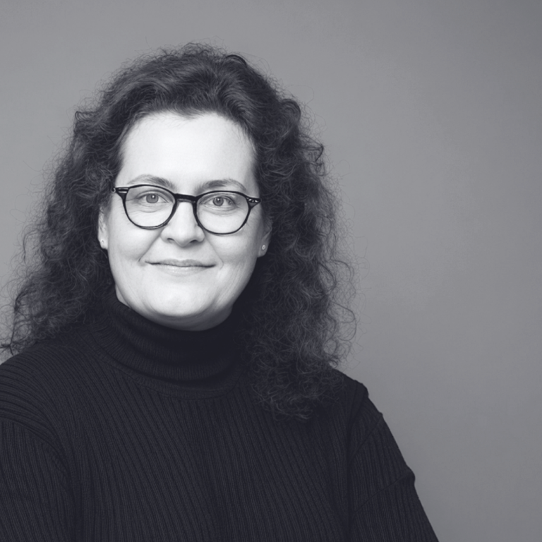 Porträt von Cornelia Schmitz. Eine freundlich lächelnde und Selbstvertrauen signalisierende Frau mit lockigem, dunklem Haar und Brille.